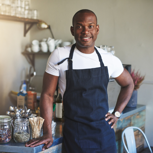 a black small business owner wearing an apron, left hand on his waist, smiling proudly